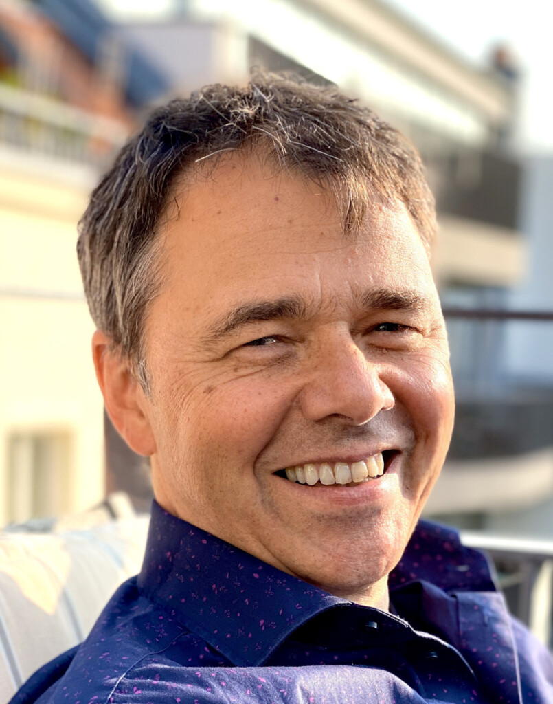 Robert Jautschus steht lächelnd auf einem Balkon, im Hintergrund sind mit Tiefenschärfe Wohnhäuser zu sehen – ein offenes Porträt, das Ruhe und Leichtigkeit vermittelt.