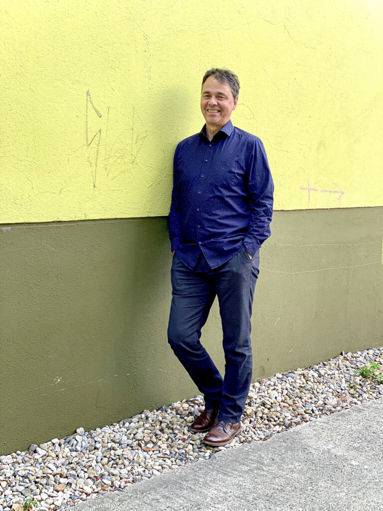 Robert Jautschus lehnt entspannt an einer Hauswand – ein ruhiges, authentisches Porträt, das Gelassenheit und Präsenz vermittelt.