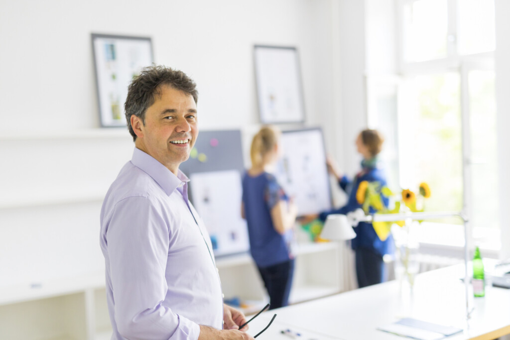 Robert Jautschus steht in einem hellen Büro, im Hintergrund unterhalten sich zwei Mitarbeiterinnen – ein authentischer Moment moderner Führung und Zusammenarbeit.